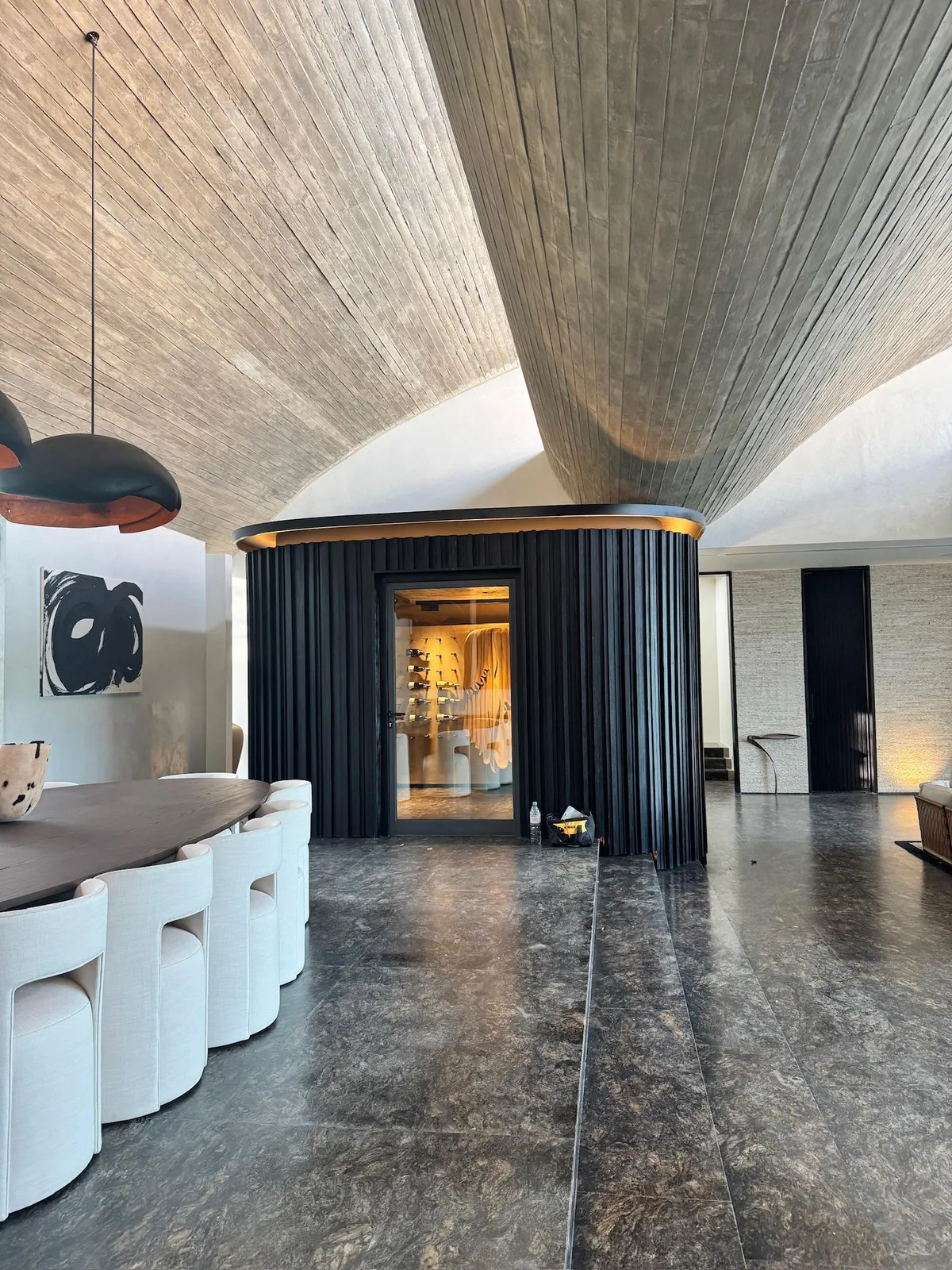 Curved black-slatted wine cellar with a glass door and warm interior lighting beneath curved wood-textured ceilings
