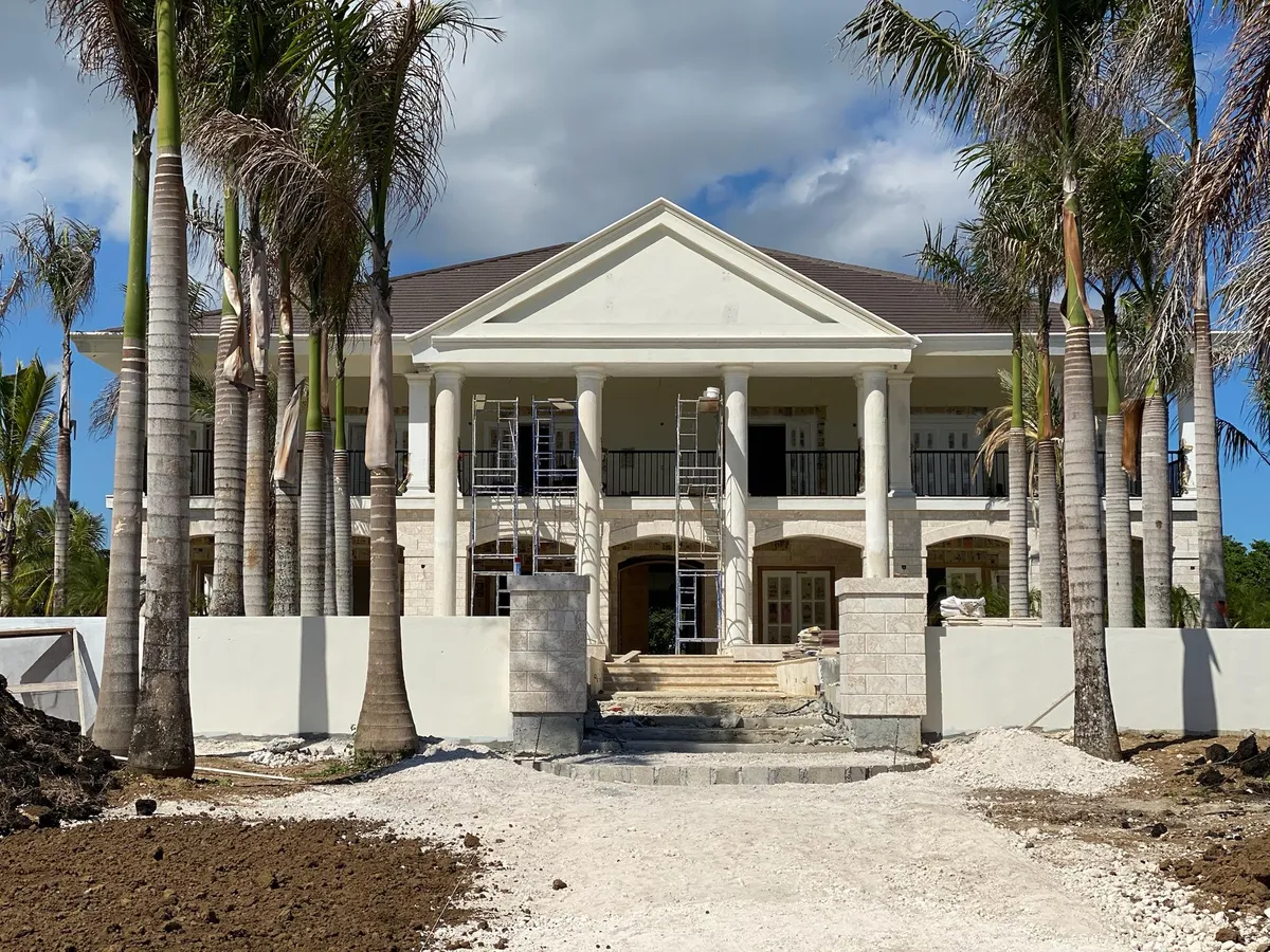 Front facade of a colonial-style mansion with a pediment and columns, lined with palms, during construction