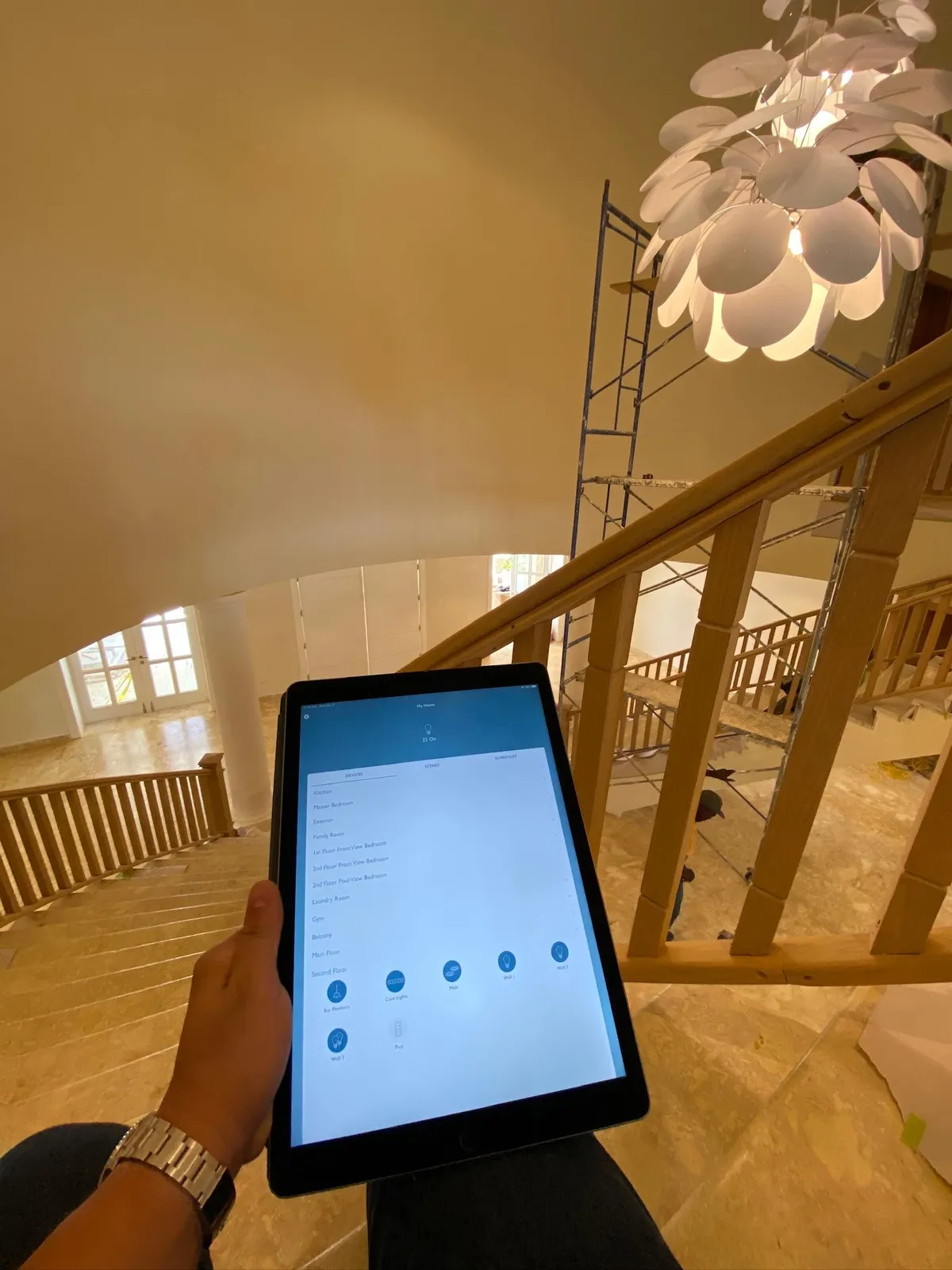 Hand holding an iPad with a lighting control app beneath a white petal pendant light over a staircase